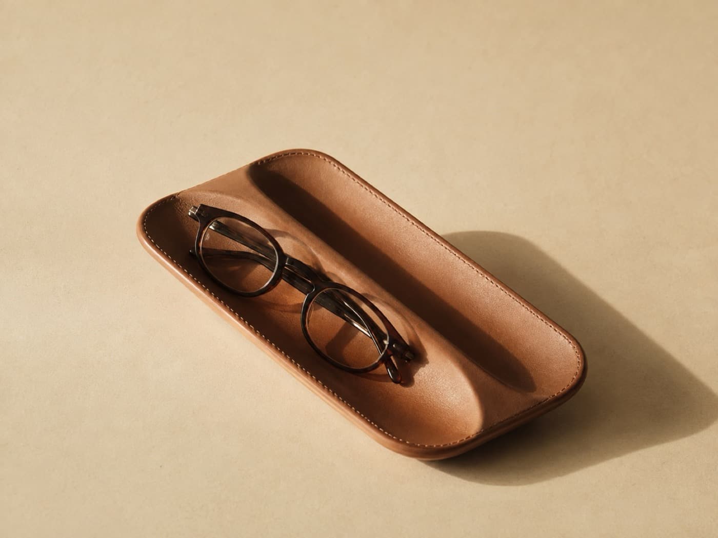 A leather caddy with two moulded channels, a pair of plain reading glasses resting in one