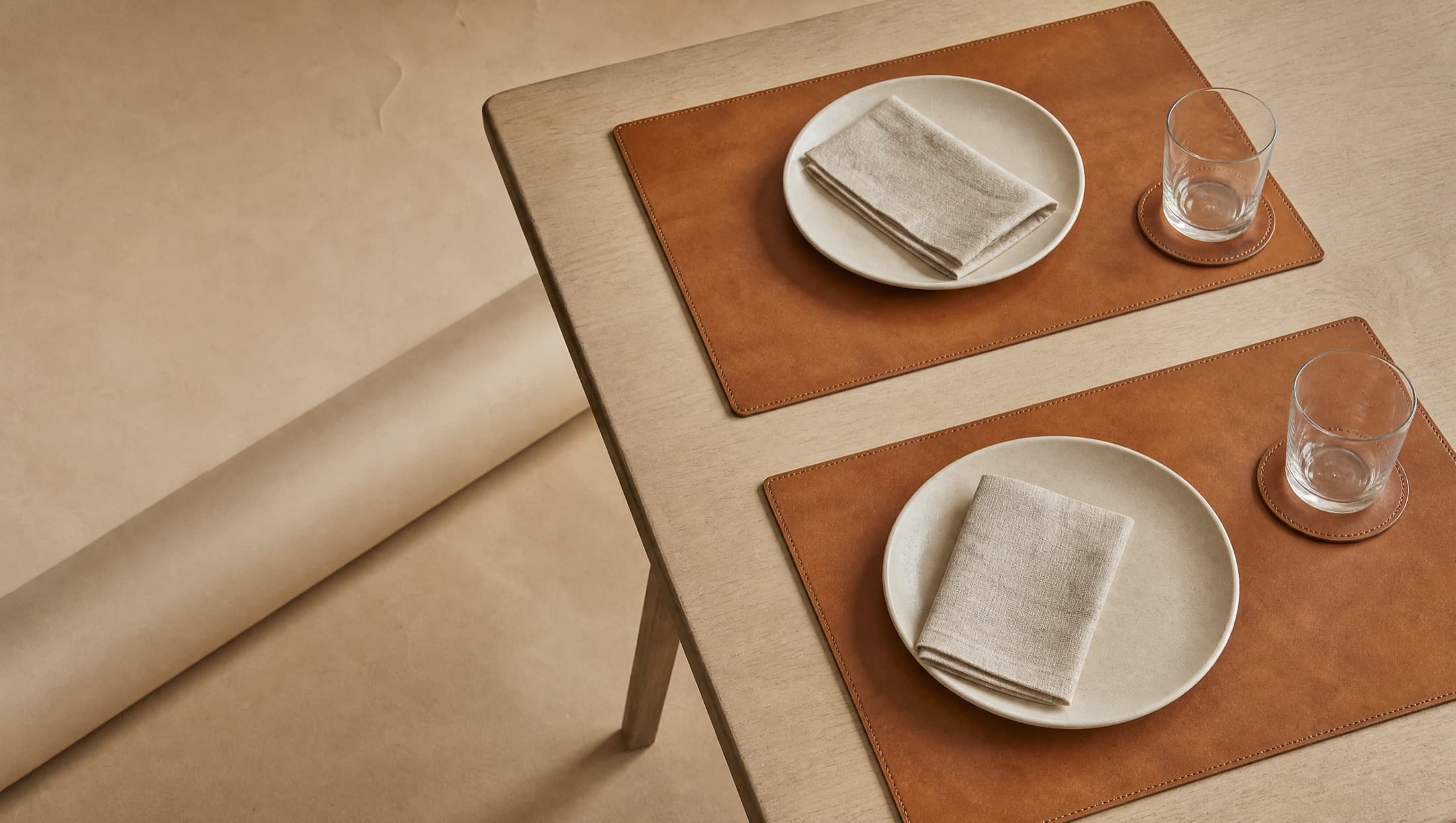 A dining table corner from above: two leather placemats with a plate and a linen napkin resting on each, and a coaster with an empty glass